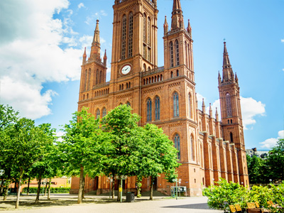 Eine rote Backsteinkirche mit mehreren Türmen und einem großen Uhrturm, umgeben von Bäumen.