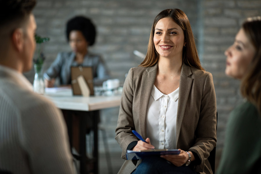 Eine Gruppe von drei Personen in einem modernen Büro. Eine Frau in einem Blazer lächelt, während sie Notizen macht. Weitere Personen sind im Hintergrund aktiv.