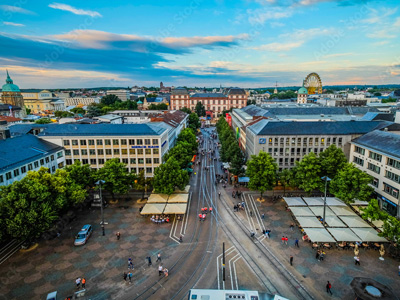 Luftaufnahme einer Stadtstraße mit grünen Bäumen, Gebäuden und einem Riesenrad im Hintergrund.