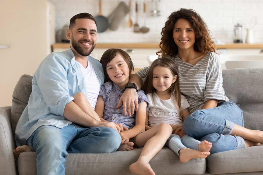Eine Familie sitzt auf einem Sofa: ein Vater, eine Mutter und zwei Kinder, ein Junge und ein Mädchen. Sie lächeln und wirken glücklich.